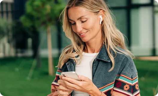 Woman with earbuds smiling while looking at her smartphone outside in a green setting.