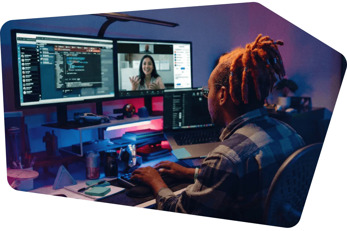 Person with dreadlocks working at a desk with two monitors, one showing code and another a video call.