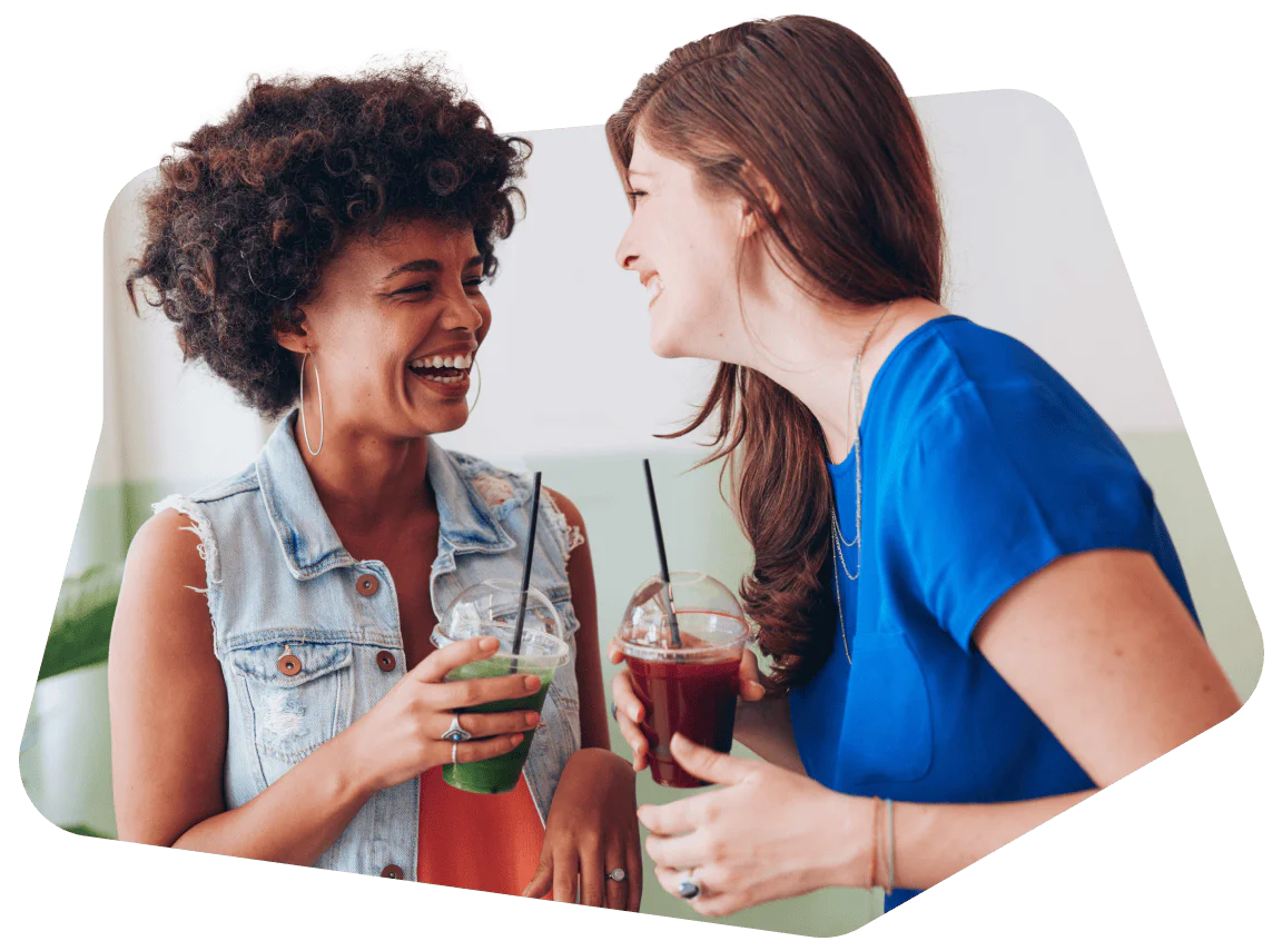 Two women smiling and holding drinks while talking inside a cafe.