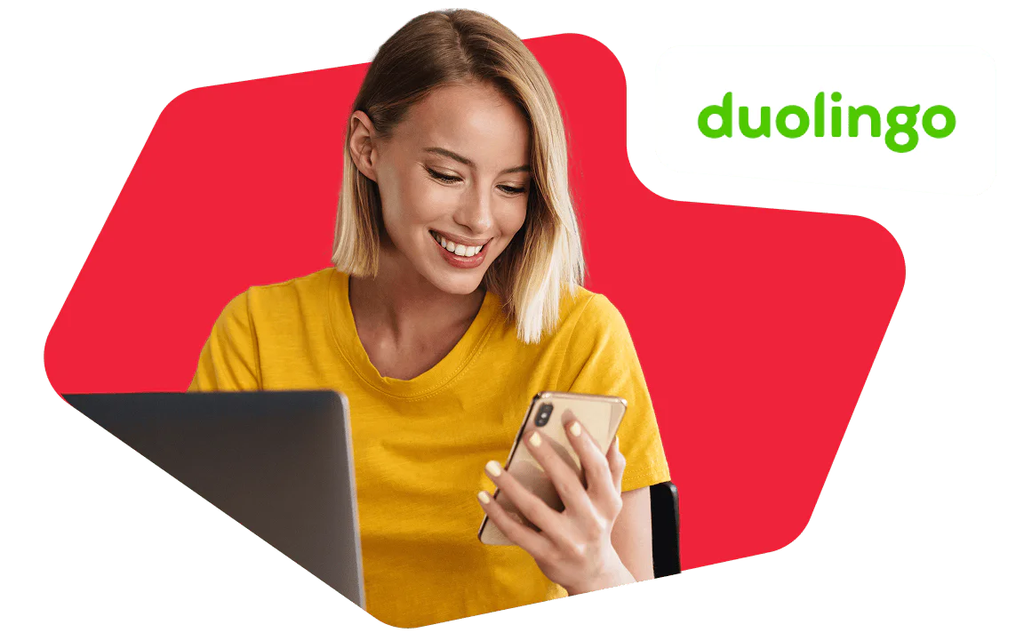 Woman Using Duolingo App on Her Phone Woman in a yellow shirt smiles at her phone with a laptop open, Duolingo logo in the corner.