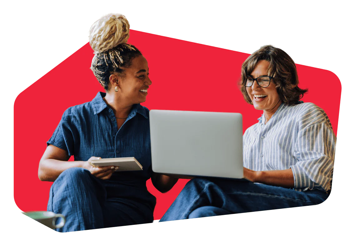 Collaborative Discussion Over Laptop Two people smiling while looking at a laptop, one holding a tablet, against a red background.