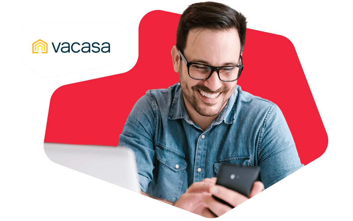 Man smiling while looking at his smartphone, with a laptop in front of him and a Vacasa logo.