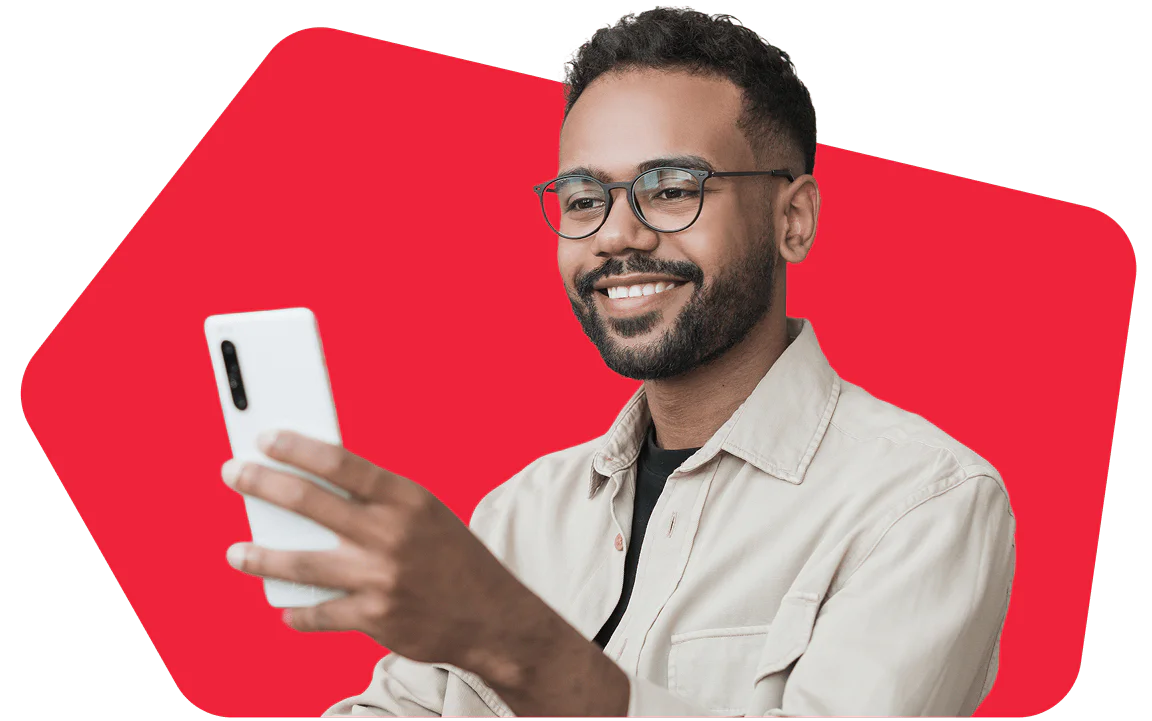 A smiling person wearing glasses and a beige shirt is holding and looking at a smartphone.