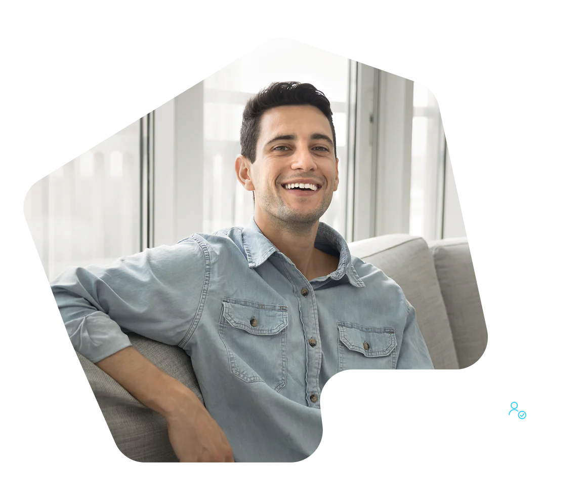 Smiling man in a light blue denim shirt sits on a gray couch in a bright room.