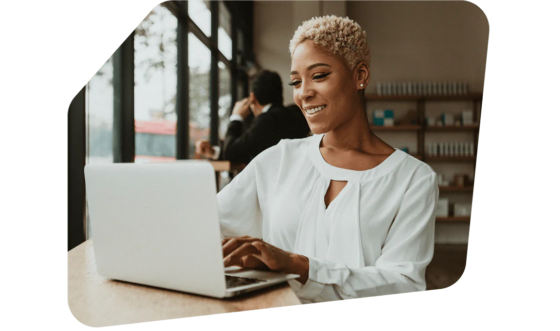 Woman smiling while using a laptop at a desk in a bright, modern workspace with colleagues in the background. Woman smiling while using a laptop at a desk in a bright, modern workspace with colleagues in the background.